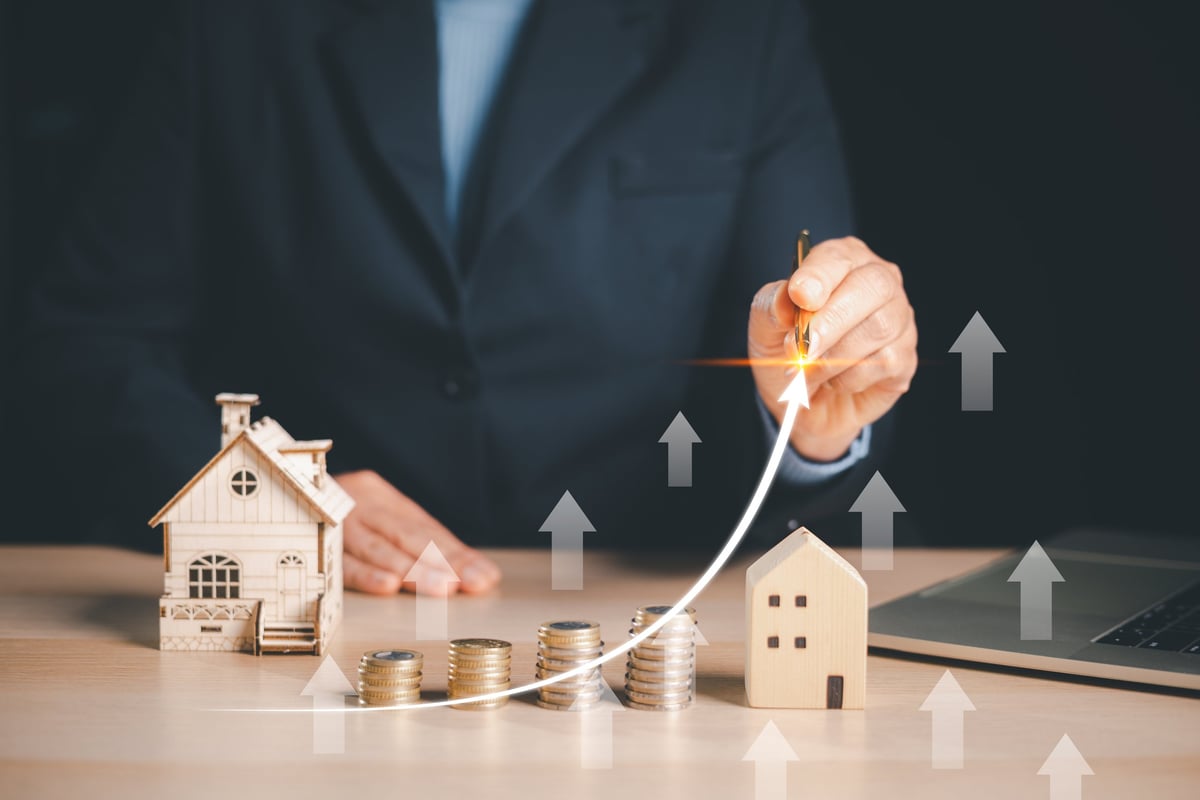 Businessman drawing rising arrow over coins, house models, showing real estate investment growth