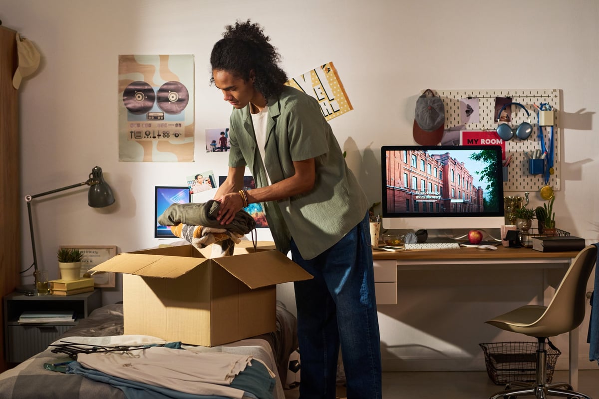 Young adult man packing folded clothes into moving box in bedroom, preparing college move