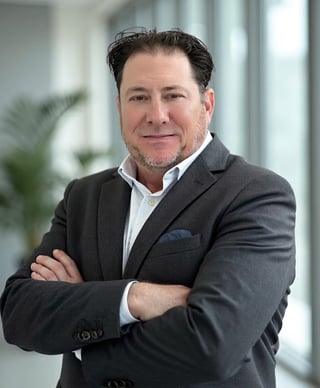 Middle-aged man in dark gray blazer and white shirt with arms crossed, smiling at camera in modern office setting